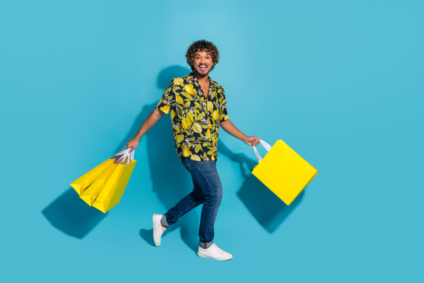 积极的男人穿着印花衬衫牛仔裤，拿着购物袋走到空旷的蓝色背景孤立的照片图片下载