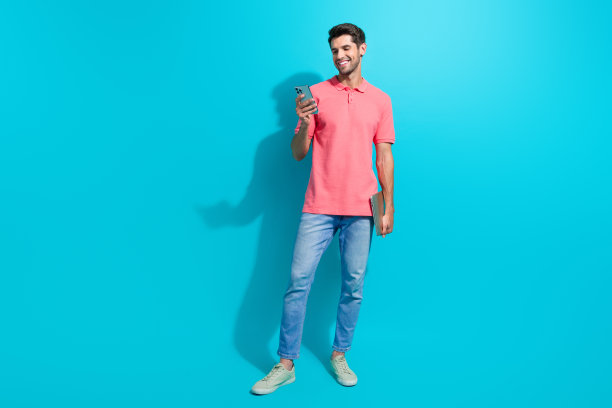 穿着粉色t恤的快乐年轻人的全身照片，使用智能手机社交网络，手持上网本孤立在青色背景上图片下载