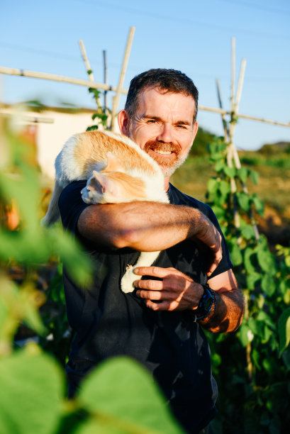 一个微笑的农夫在他的肩膀上抱着一只猫在他的乡村花园图片下载