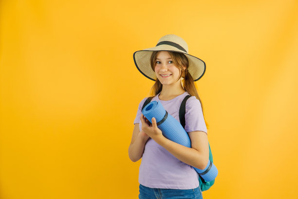 快乐可爱的小女孩旅行者背着徒步背包，徒步旅行，站在黄色的背景上，戴着时髦的帽子，脸上带着快乐的表情，爱旅行图片下载