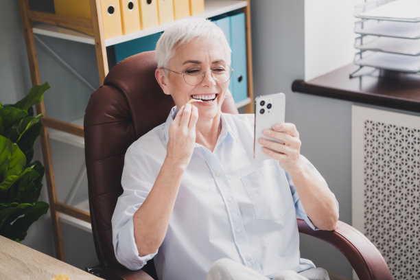 快乐迷人的年长女秘书穿着正装衬衫，吃着薯片，在室内车间工作站交流现代设备的照片图片下载