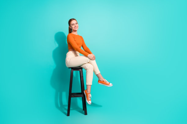 成功女性的全身照片，穿着露臀裤，坐在酒吧椅子上，双腿交叉，在绿松石色的背景上分开图片下载