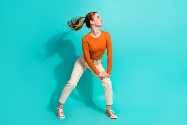 全身照片可爱的女人飘飘的头发，穿露脐上衣白色裤子，手臂放在膝盖上孤立的绿松石色背景图片下载