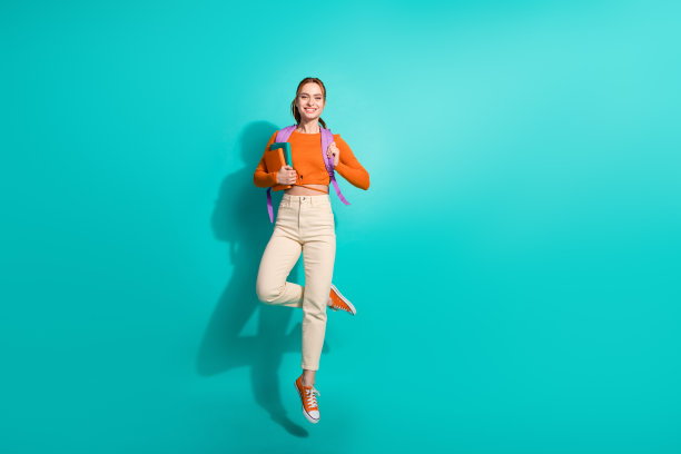 欢快的女人穿着露脐裤的全身照片，肩上背着背包，在绿松石色的背景上抱着书飞图片下载