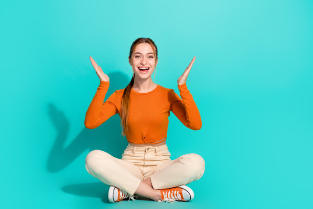 印象深刻的女子全身照片穿着露脐白裤坐在地板上举起手掌孤立的绿松石色背景图片下载