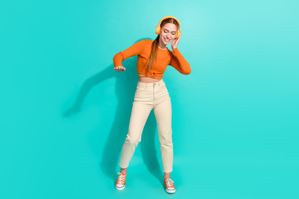 心情好女孩的全尺寸照片穿着橙色衬衫裤子触摸耳机享受立体声孤立的蓝绿色背景图片下载
