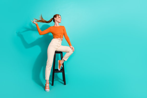 漂亮女孩的全尺寸照片，穿着橙色衬衫，坐在凳子上，飘动着尾巴，在蓝绿色的背景上看起来空荡荡的图片下载