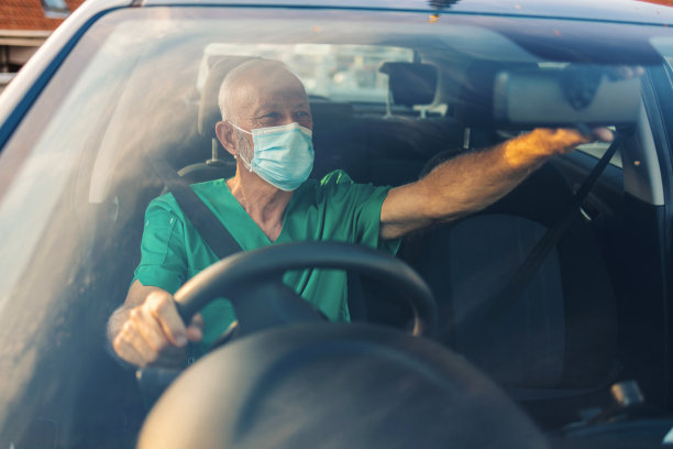 白人男医生在开车。成熟的微笑护士下班后开车回家。白天开车时，戴着医用服和口罩的自信的医务人员。男性医疗保健专业人员开车上班。图片下载