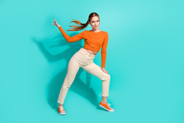 花式女子的全身照片与飘飘的头发穿橙色上衣裤手在腿上孤立的绿松石色背景图片下载