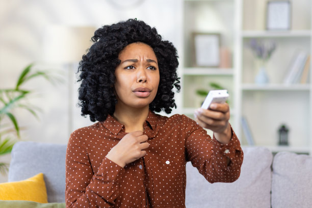 一位年轻的非裔美国女性用遥控器打开空调的特写照片，坐在家里的沙发上，感觉不舒服，喘不过气来图片下载
