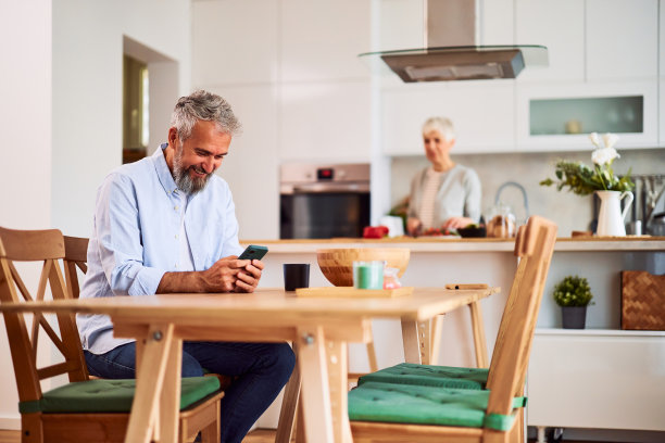 一位年长的成年男子坐在餐桌旁，一边耐心地等待妻子在厨房准备的早餐，一边用手机发短信图片下载