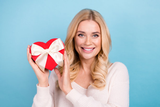 照片中迷人可爱的女人穿着时髦的白色衣服，拿着红色的礼盒，孤立在蓝色的背景上图片下载