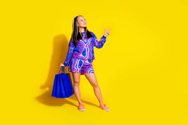 漂亮女人穿着套头衫，拿着购物袋的全长照片，看着黄色背景上的标志空白空间图片下载