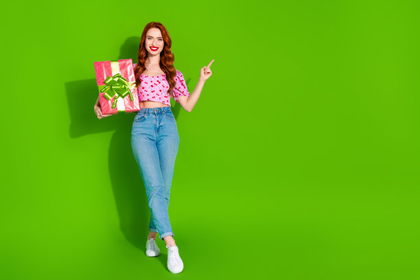 全身照片光泽迷人的女士穿着印花顶持有礼盒指向空白空间孤立的绿色背景图片下载