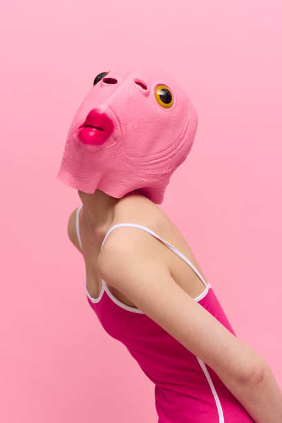 一个瘦削的女人穿着粉红色的万圣节服装，脸上有一个鱼头，在粉红色的背景下滑稽地摆姿势，看着相机，艺术概念照片图片下载