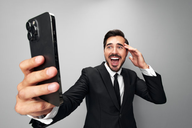 男子商人手里拿着手机惊讶地看着屏幕上的视频通话和自拍，笑得龇牙咧嘴。特写广角照片灰色孤立背景图片下载