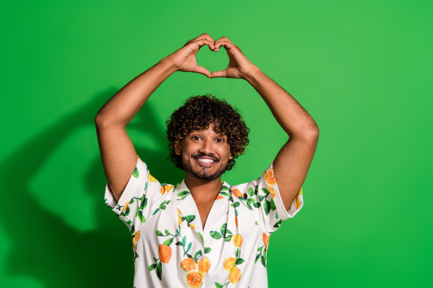 照片中，开朗兴奋的阿拉伯男子穿着印花衬衫，手臂上升，露出心形手势，孤立的绿色背景图片下载