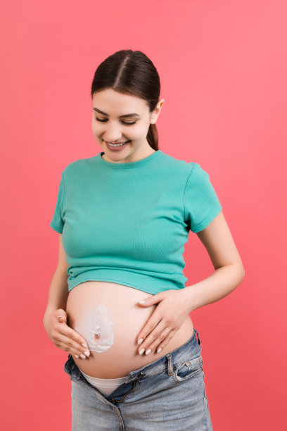 穿着敞开牛仔裤的孕妇在腹部涂抹保湿乳液，以避免在彩色背景和复制空间上出现妊娠纹。孕期护肤概念图片下载
