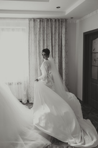 一个美丽的黑发新娘正准备在一个美丽的闺房风格的衣服走到她的婚礼礼服的窗口。黑白照片。图片下载