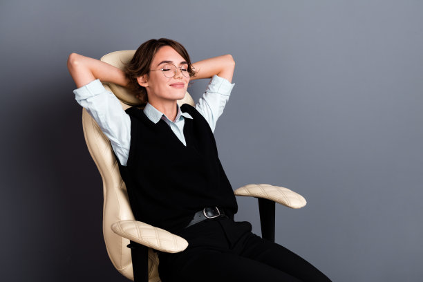 照片中满意的女人，梳着鲍勃发型，戴着眼镜，穿着正装，躺在扶手椅的手臂上，头后面是灰色的背景图片下载