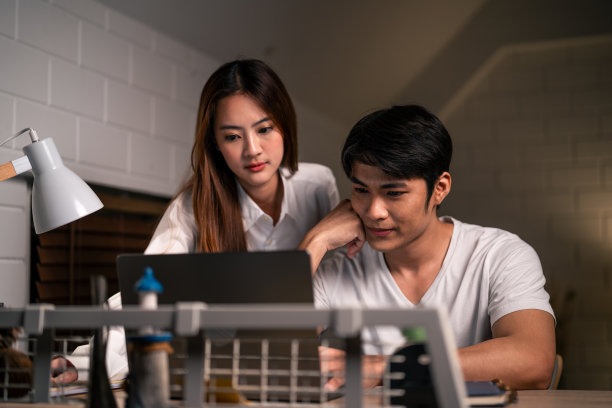 亚洲年轻人晚上在家和妻子在卧室里加班。迷人美丽的妻子穿着睡衣等着丈夫用笔记本电脑工作，在家里睡觉时上网交流。图片下载