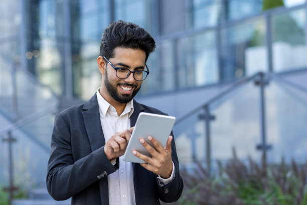 特写照片，一个微笑的年轻印度男子穿着西装坐在办公大楼附近使用平板电脑图片下载