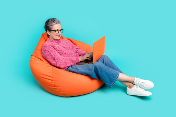 聪明的人穿着针织套头衫的全尺寸照片，坐在沙发上，看着笔记本电脑，在绿松石色的背景上工作图片下载