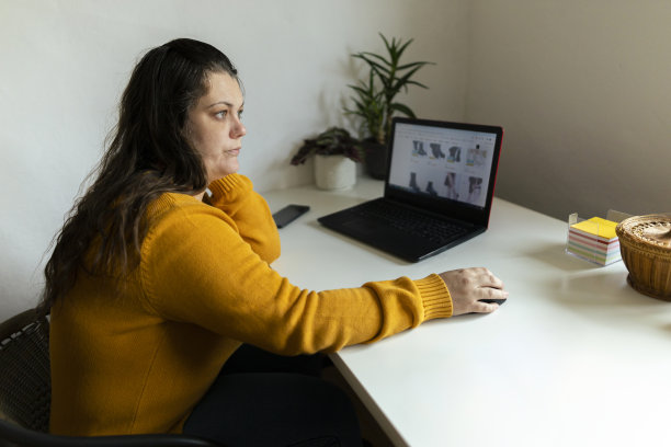 网上购物在家里，美丽的女人使用笔记本电脑在她的家庭工作空间和网上购物。穿着舒适的黄色毛衣。库存图片图片下载