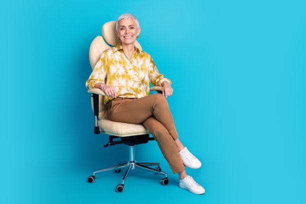 漂亮退休女性的全尺寸照片，坐在办公室扶手椅上，穿着时髦优雅的衣服，孤立在蓝色背景上图片下载