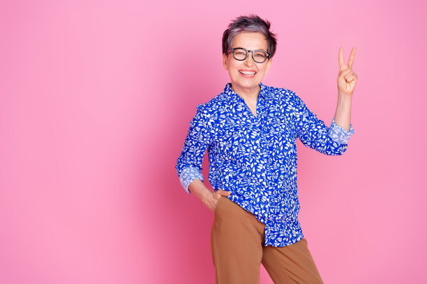 照片积极的人与灰色的发型穿着印花衬衫hold手臂在口袋里显示v字孤立的粉红色背景图片下载