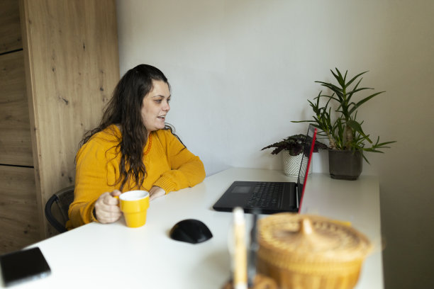 网上购物在家里，美丽的女人使用笔记本电脑在她的家庭工作空间和网上购物。穿着舒适的黄色毛衣。库存图片图片下载