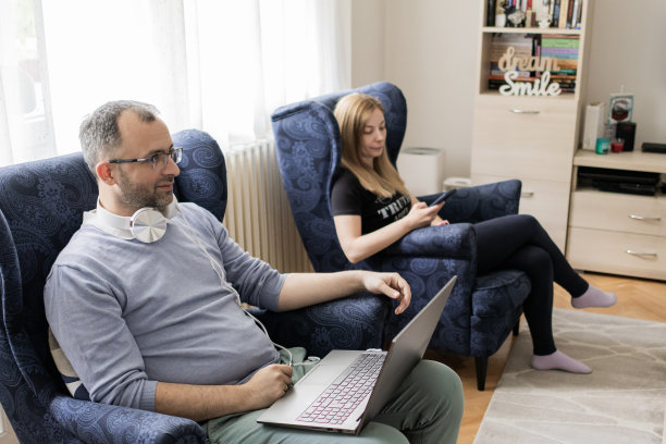 现代夫妻一起在家。男人正在用笔记本电脑工作，而他的妻子坐在他旁边用手机。感觉工作和家庭生活平衡和谐的人。图片下载