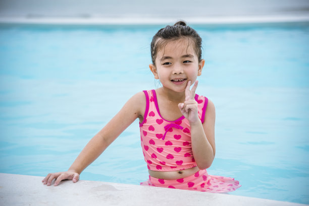照片中的女孩在粉红色的泳衣在游泳池嬉戏夏天时间与粉红色的泳衣，蓝色的水在酒店游泳池户外。蹒跚学步的女孩孩子笑笑看着镜头。快乐女孩游泳嬉戏图片下载