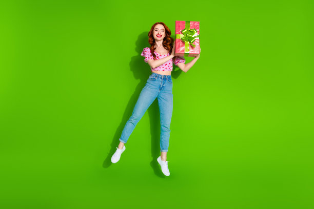 照片中迷人的正面女子穿着印花上衣，手持礼盒跳高，空旷的空间孤立绿色背景图片下载