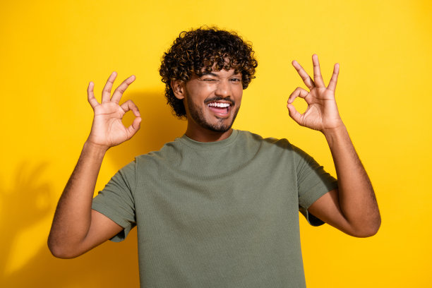 照片中，一个穿着卡其色t恤，有着波浪发型的时髦男人，在黄色的背景下，手指上露出眨着的眼睛图片下载