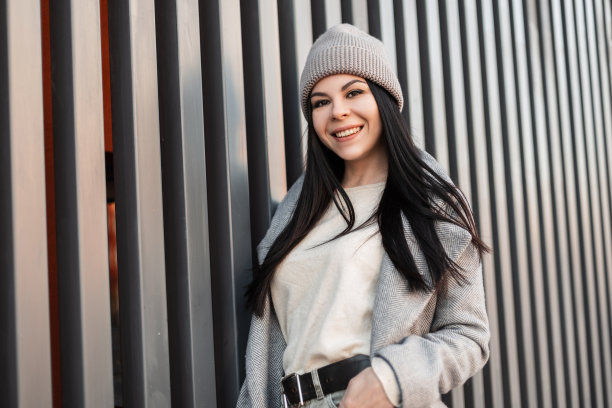 照片中，年轻的黑发女子穿着时尚的春季外套，带着可爱的微笑，在这座城市的现代灰色墙壁旁摆姿势。快乐的女孩在户外微笑。时髦的女性服装。街头风格。图片下载