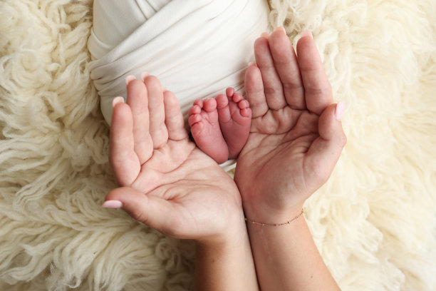 新生儿的脚放在父母的手掌上。父亲和母亲的手掌托着新生婴儿的脚，上面盖着一条白色的弗洛卡蒂毯子。孩子的脚趾、脚后跟和脚的照片。图片下载