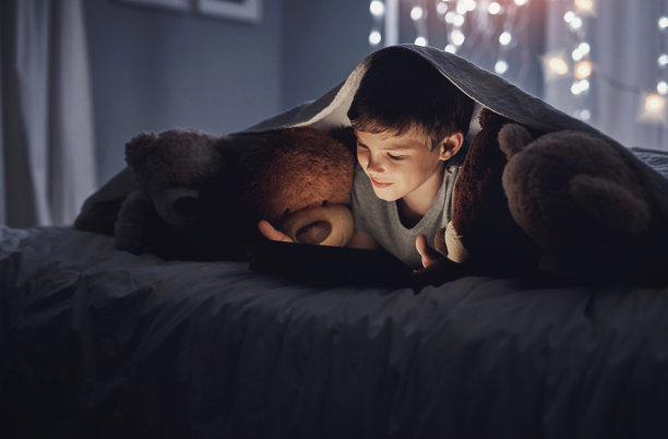 晚上，男孩、孩子和平板电脑躺在床上看动画片、玩网络游戏或盖着毯子看电影。孩子，泰迪熊和数字技术电子书，视频订阅和讲故事的应用程序在家里图片下载