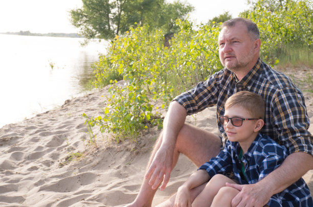 父亲节概念。父亲和儿子一起坐在河边，望着远方。图片下载