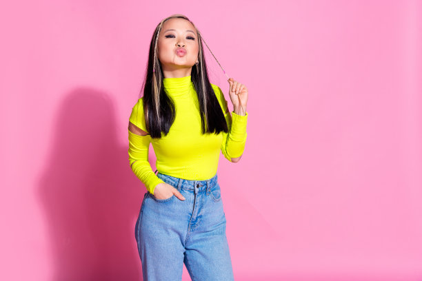 照片中的漂亮女孩染了头发，穿着黄色高领毛衣，手臂插在口袋里，拿着辫子，在粉红色的背景上孤立地飞吻图片下载