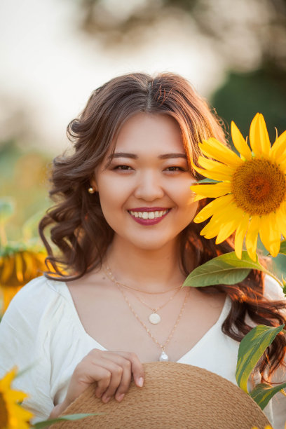 日落时分，年轻的亚洲卷发女子站在向日葵地里。一个年轻美丽的亚洲女人在阳光下的肖像。图片下载