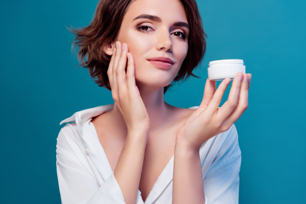 新鲜皮肤的特写照片，年轻女士拿着一瓶乳液，涂在脸上，丝滑的皮肤被隔离在蓝色背景上图片下载