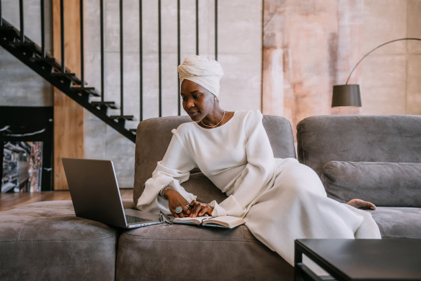 一名身穿白色套装、头裹头巾的黑人女性坐在沙发上用笔记本电脑工作，在一张特写照片中，她微笑着，在另一张照片中，她抱着一只白色的小狗图片下载