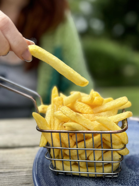 照片中，一个面目全非的红发女人从盘子里装着炸薯条的金属篮子里拿起薯条，用食指和拇指夹着薯条，露天木质餐桌，背景是鸡肉汉堡和薯条图片下载