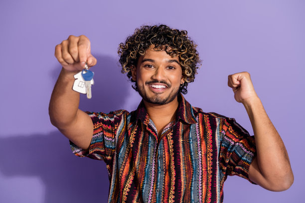 照片上满意的男人留着胡子，卷发，穿着印花衬衫，手臂拿着新房的钥匙，孤立在紫色的背景上图片下载