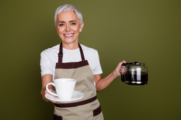 照片中可爱快乐的女人穿着围裙，端着饮料，给水壶上水，空旷的空间孤立的卡其色背景图片下载