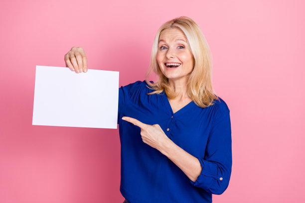 照片肖像迷人的退休妇女拿点激动的白纸床单穿着蓝色的衣服孤立在粉红色的背景图片下载