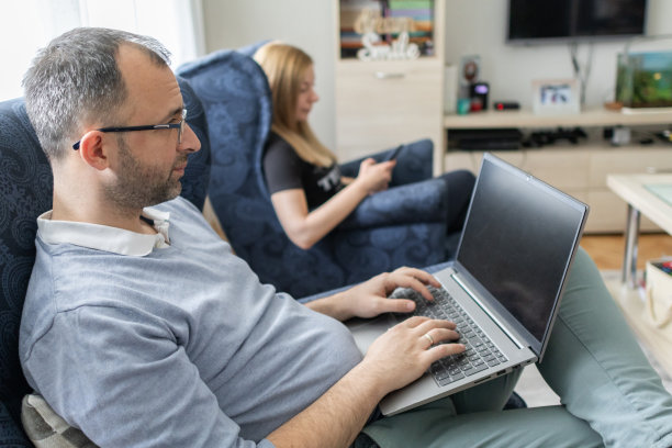 现代夫妻一起在家。男人正在用笔记本电脑工作，而他的妻子坐在他旁边用手机。感觉工作和家庭生活平衡和谐的人。图片下载