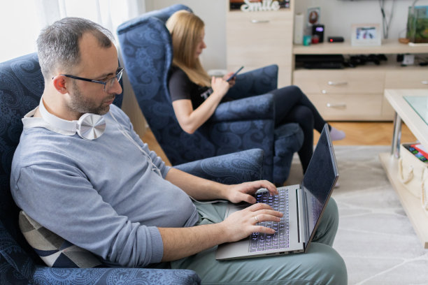 现代夫妻一起在家。男人正在用笔记本电脑工作，而他的妻子坐在他旁边用手机。感觉工作和家庭生活平衡和谐的人。图片下载