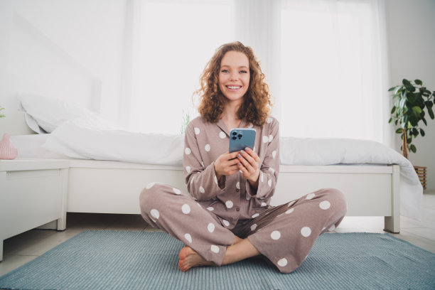 漂亮的年轻女孩的全尺寸照片使用智能手机穿着睡衣明亮的室内卧室公寓室内图片下载